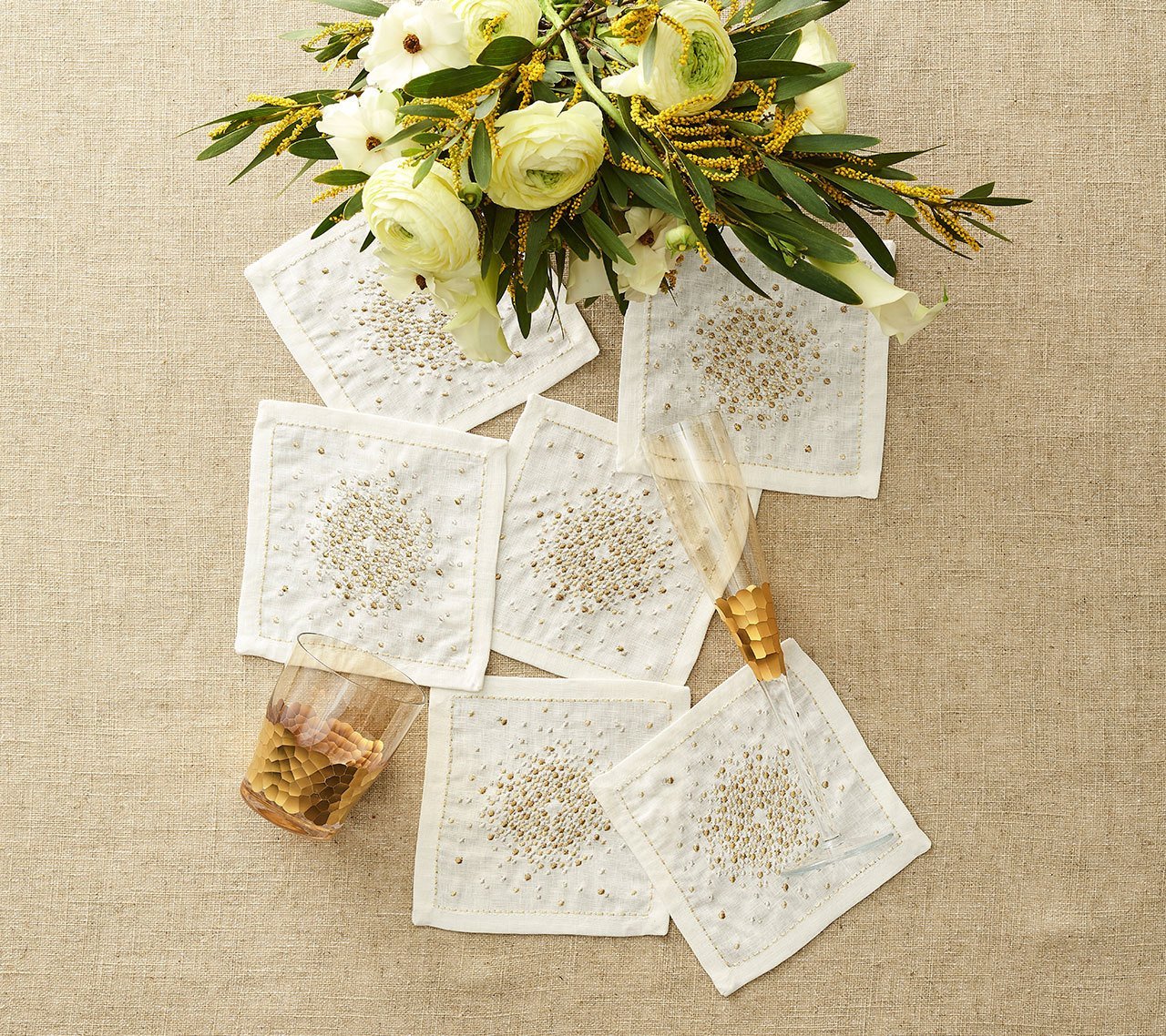  Luxury Starburst Cocktail Napkins in white, gold & silver, shown with a floral arrangement and gold wine glasses