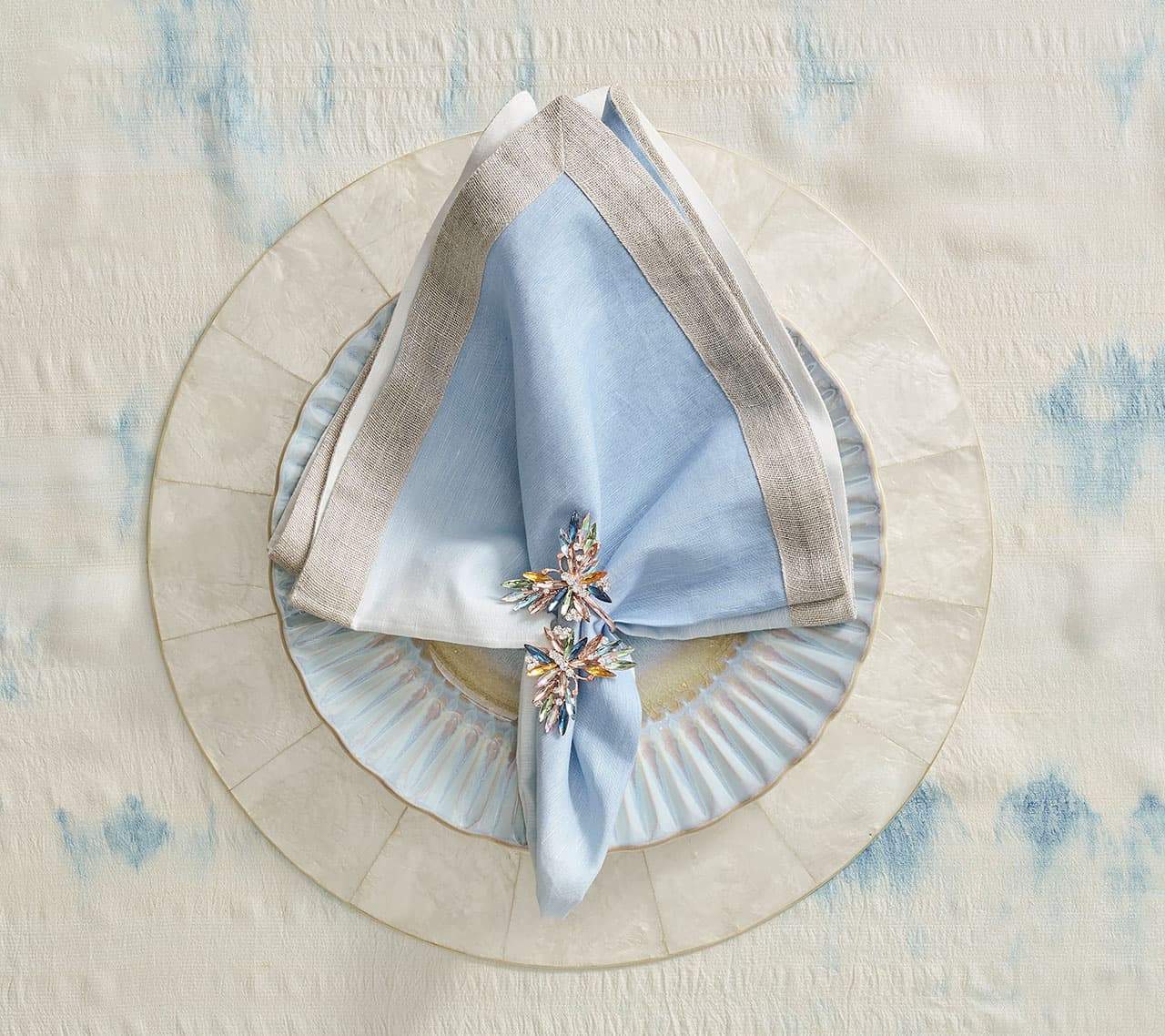 Jeweled napkin ring holding a blue and white linen napkin on top of the round Capiz Placemat in Natural