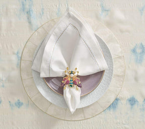 Jeweled fly napkin ring holding a white napkin on a purple plate underneath a round Capiz Placemat in natural