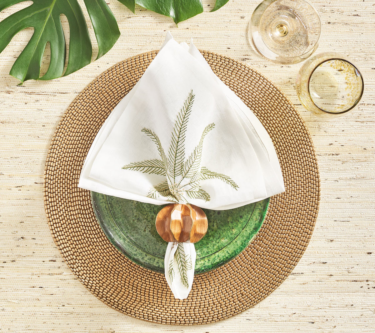 Place setting with a brown, round placemat below a whitePalm Coast Napkin with a green & gold palm leaf