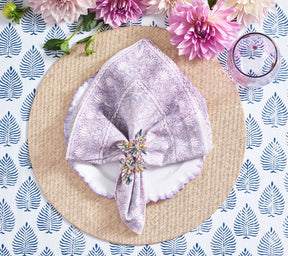 Natural and gold Glam Grass Placemat, lavender-rimmed plates, lavender napkin, pink flowers on a blue & white tablecloth