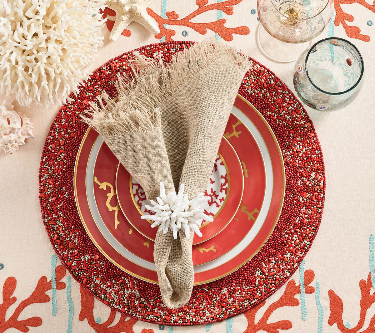 Place setting with coral plates, a natural napkin held by a white napkin ring and a coral & gold beaded Maui Placemat 