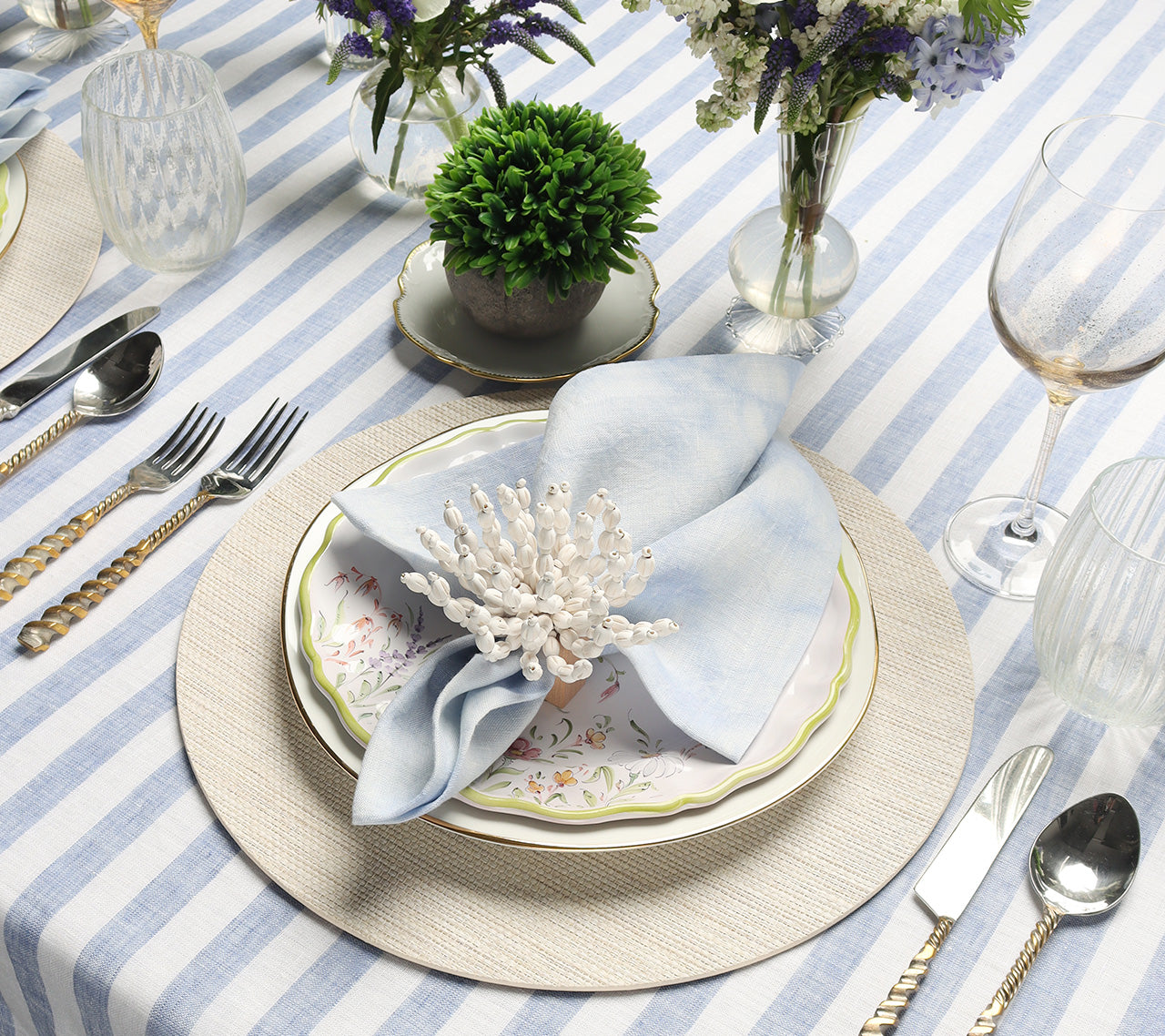 White & blue striped tablecloth,  gold cutlery, Seaside Placemat in natural, white floral plate, blue napkin and flowers in clear vases