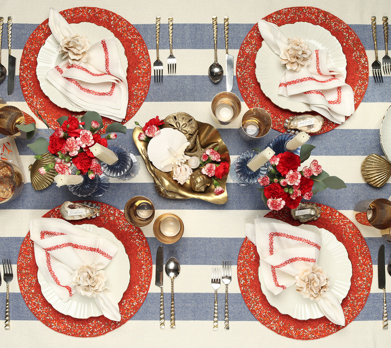 Four place settings with coral & gold beaded Maui Placemats, white plates, white napkins with a coral border on top of a blue & white stripe tablecloth