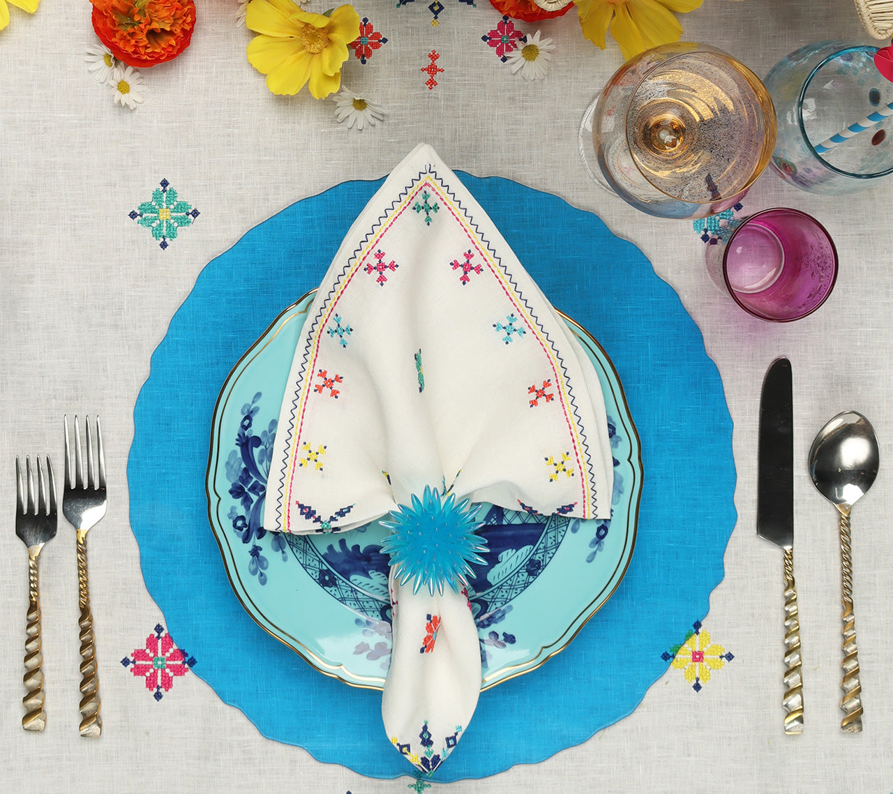 Place setting with a blue placemat and a Kim Seybert Luxury Fez Napkin in white & multi